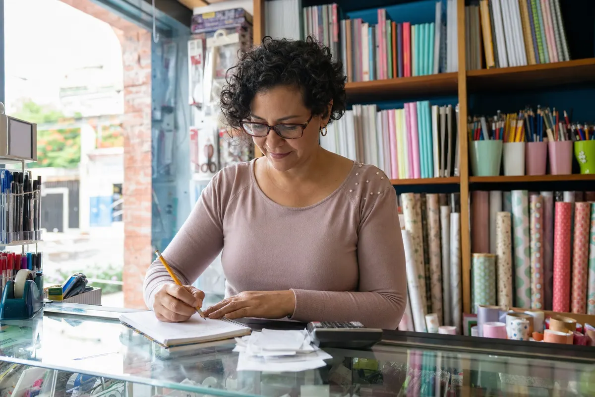 Dueña de un negocio pequeño anotando ingresos y gastos en su mesa de trabajo con el celular y recibos